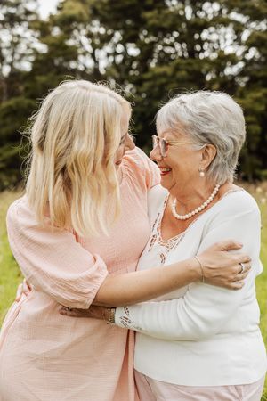 Two women embracing and laughing outdoors, one blonde in pink and one elderly with gray hair in white, sharing a joyful moment.