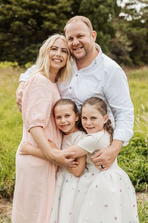 Happy family of four posing outdoors, parents with two young daughters in white dresses, smiling in a green park setting.