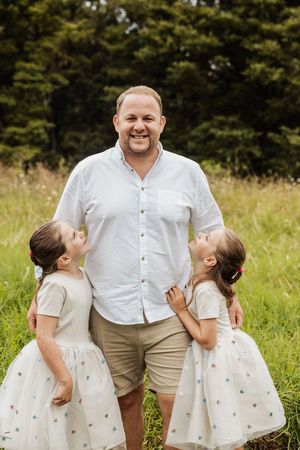 A smiling man in a white shirt stands in a green field with two young girls in matching white dresses looking up at him.