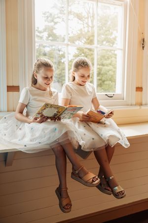 Two young girls in white tulle dresses sit on a windowsill reading books together in a bright, cozy room.