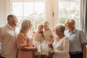 Multi-generational family of six smiling together indoors near a bright window in a warm, candid portrait.