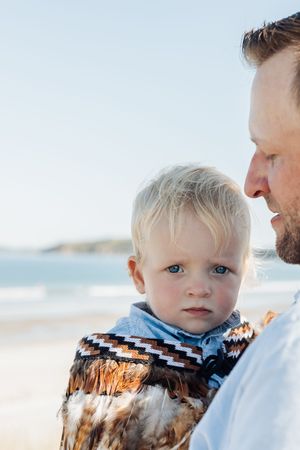 Father holding blond toddler with blue eyes on a sunny beach, child wearing a feathered wrap.