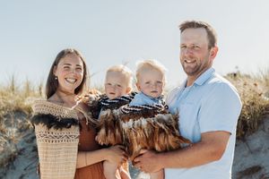 Happy family of four posing on a sunny beach, parents holding two young toddlers near sand dunes.