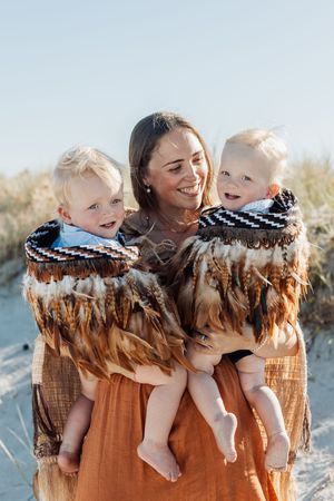 Smiling mother holding twin toddlers wrapped in a traditional Maori feather cloak on a sunny beach.