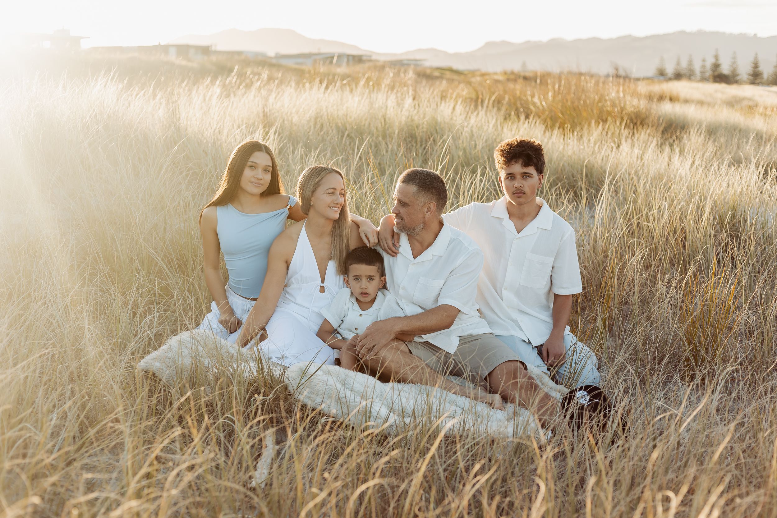 Family of five sitting together in golden grass field during sunset, wearing white and light blue outfits.