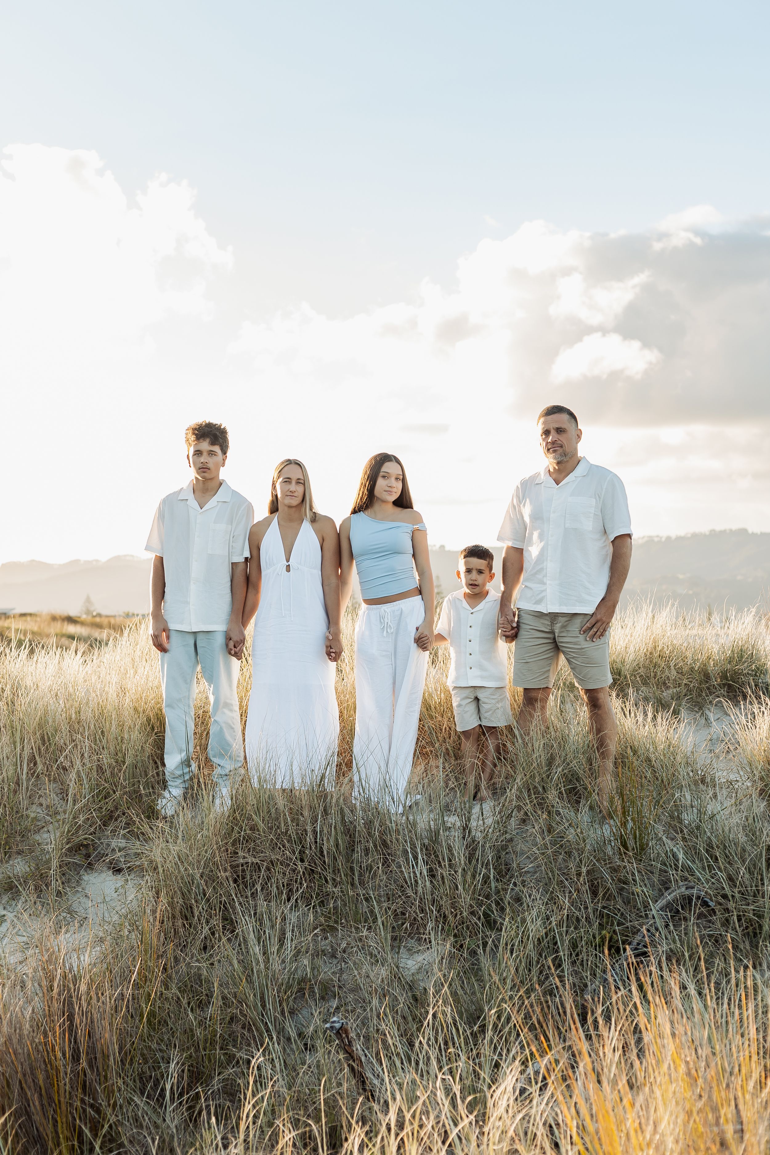 Family of five posing together in golden coastal dunes, dressed in white and light blue outfits at sunset.