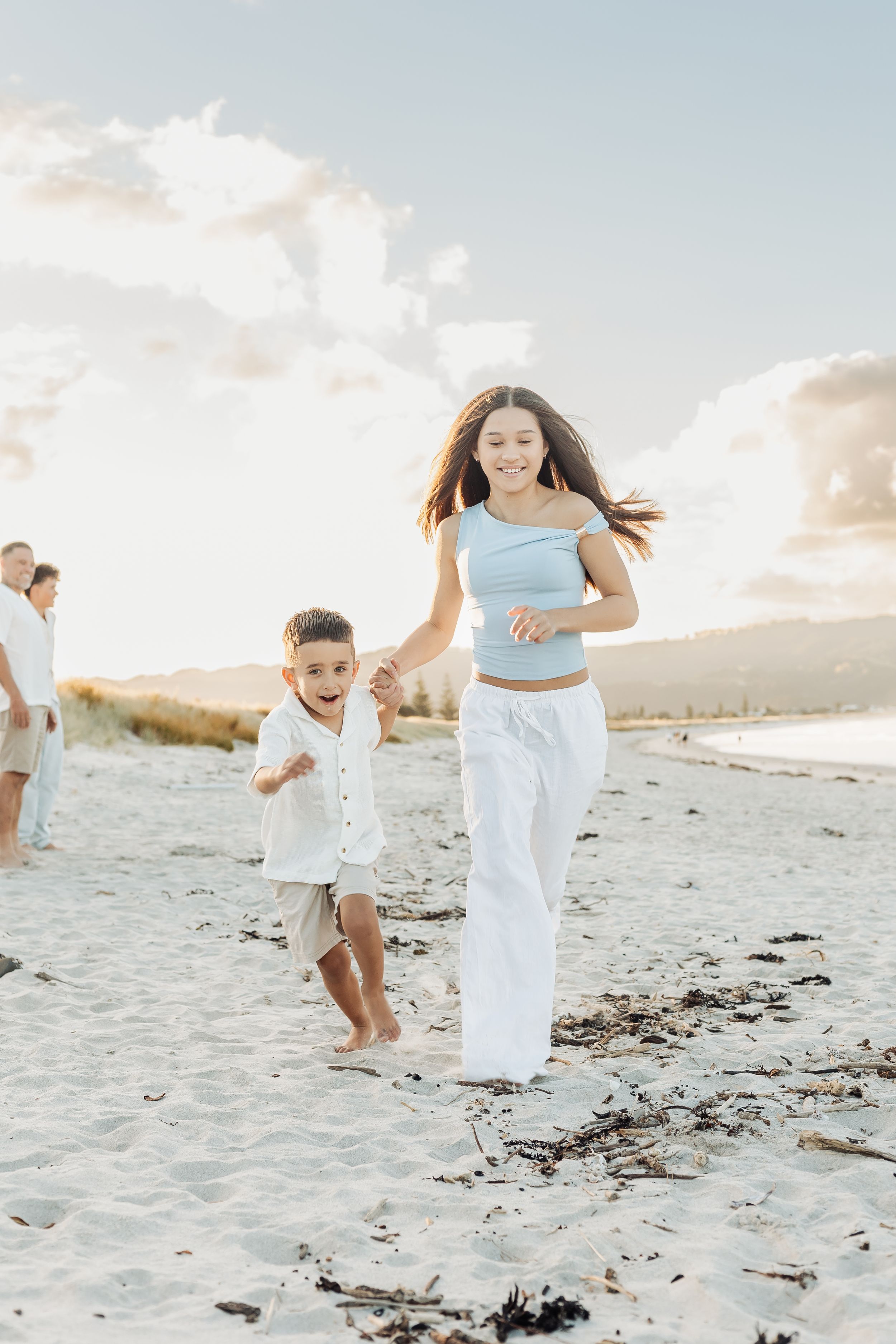 A girl and young boy run joyfully hand-in-hand on a sandy beach at sunset, dressed in white and blue.