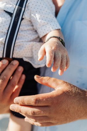 Close-up of an adult hand gently supporting a sandy baby hand at the beach, with a bracelet visible.