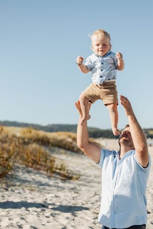 Father joyfully lifting baby boy in the air on a sunny sandy beach, both wearing casual blue shirts.
