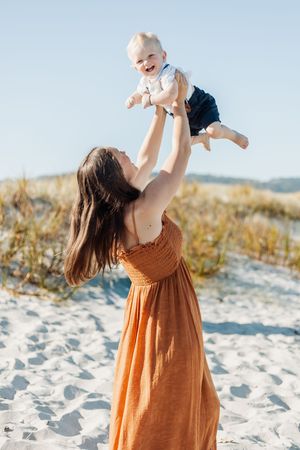 A joyful mother in an orange dress lifts her laughing baby high in the air on a sunny sandy beach.