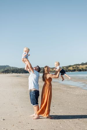 A happy family of four plays on a sunny beach, parents lifting two toddlers in the air near the ocean.
