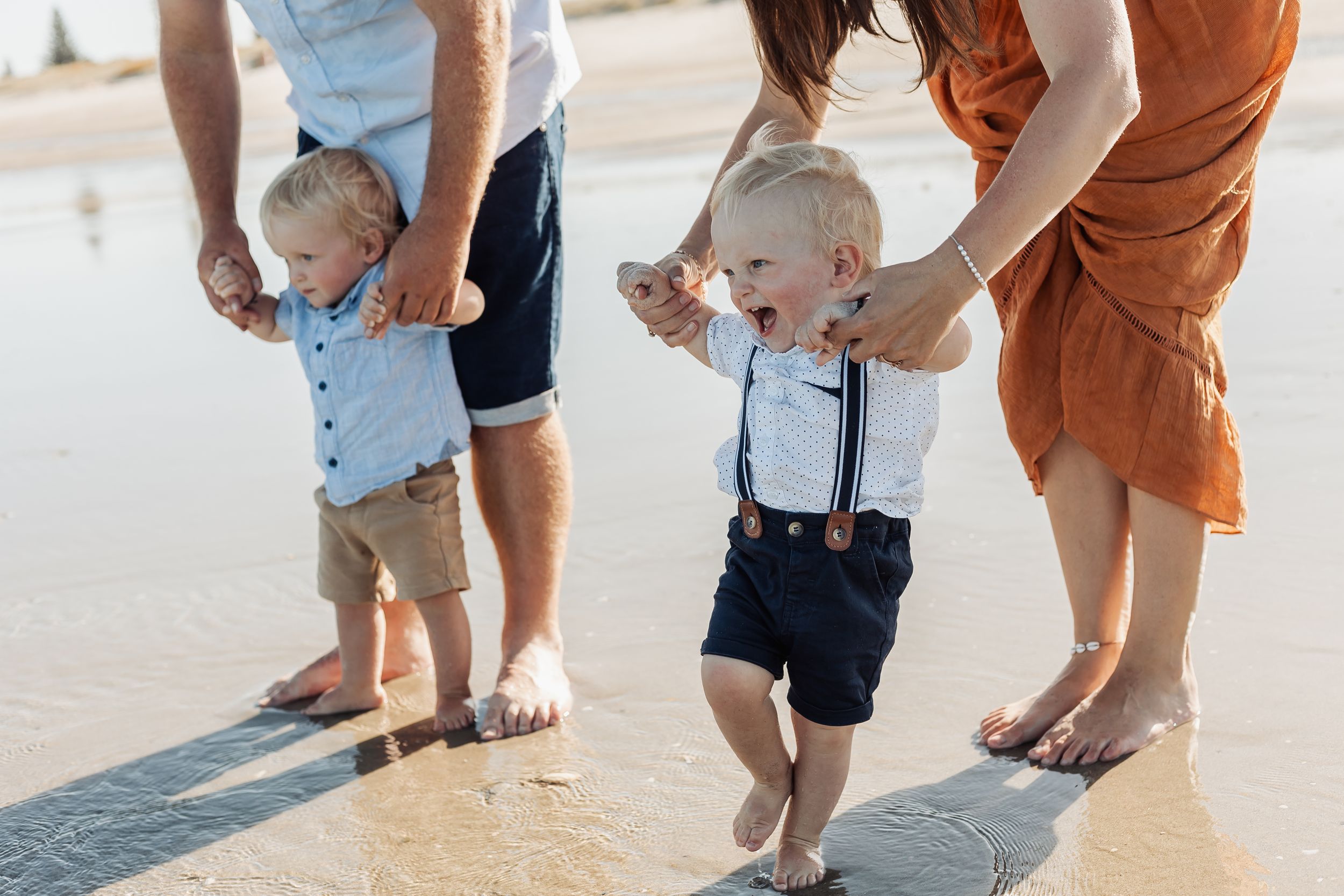 Two toddlers learning to walk on a sunny beach, supported by their parents hands, dressed in cute matching outfits.