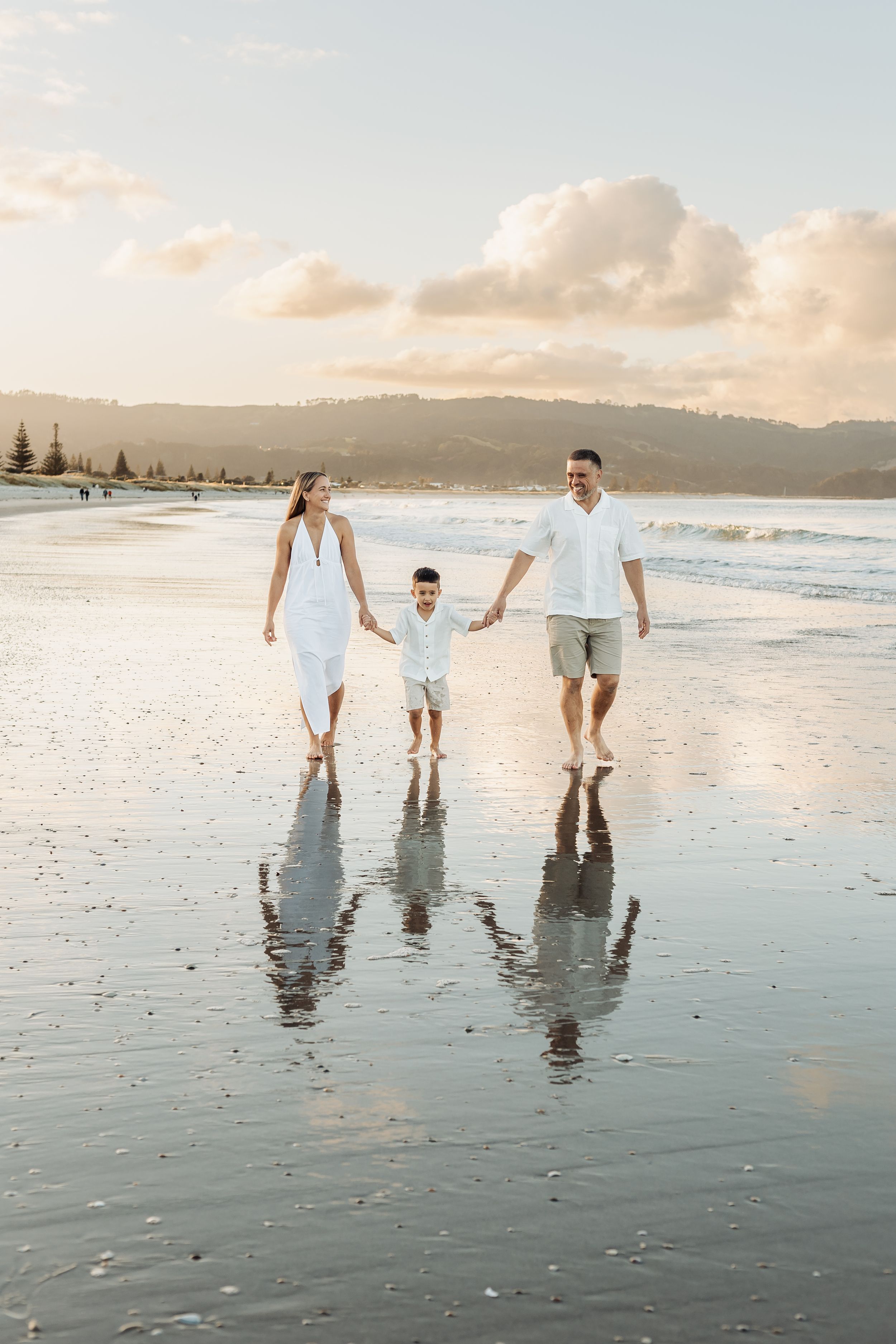 Family of three walking hand-in-hand on a reflective beach at sunset, parents in white clothing with young child between them.