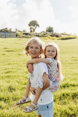 Two blonde girls smile outdoors in a sunny green field, one giving the other a piggyback ride.