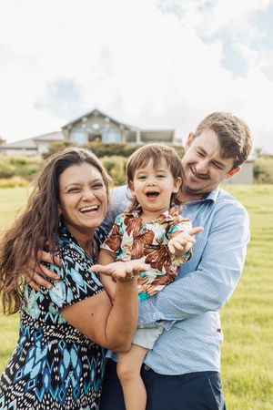 Happy family of three laughing outdoors, parents holding excited toddler in floral shirt on green lawn.