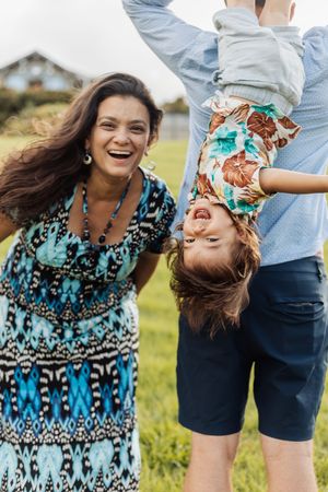 A joyful family outdoors, mother laughing as father holds child upside down on green grass.