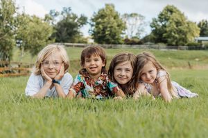 Four smiling children lying on green grass in a sunny park, posing together for a cheerful outdoor family photo.