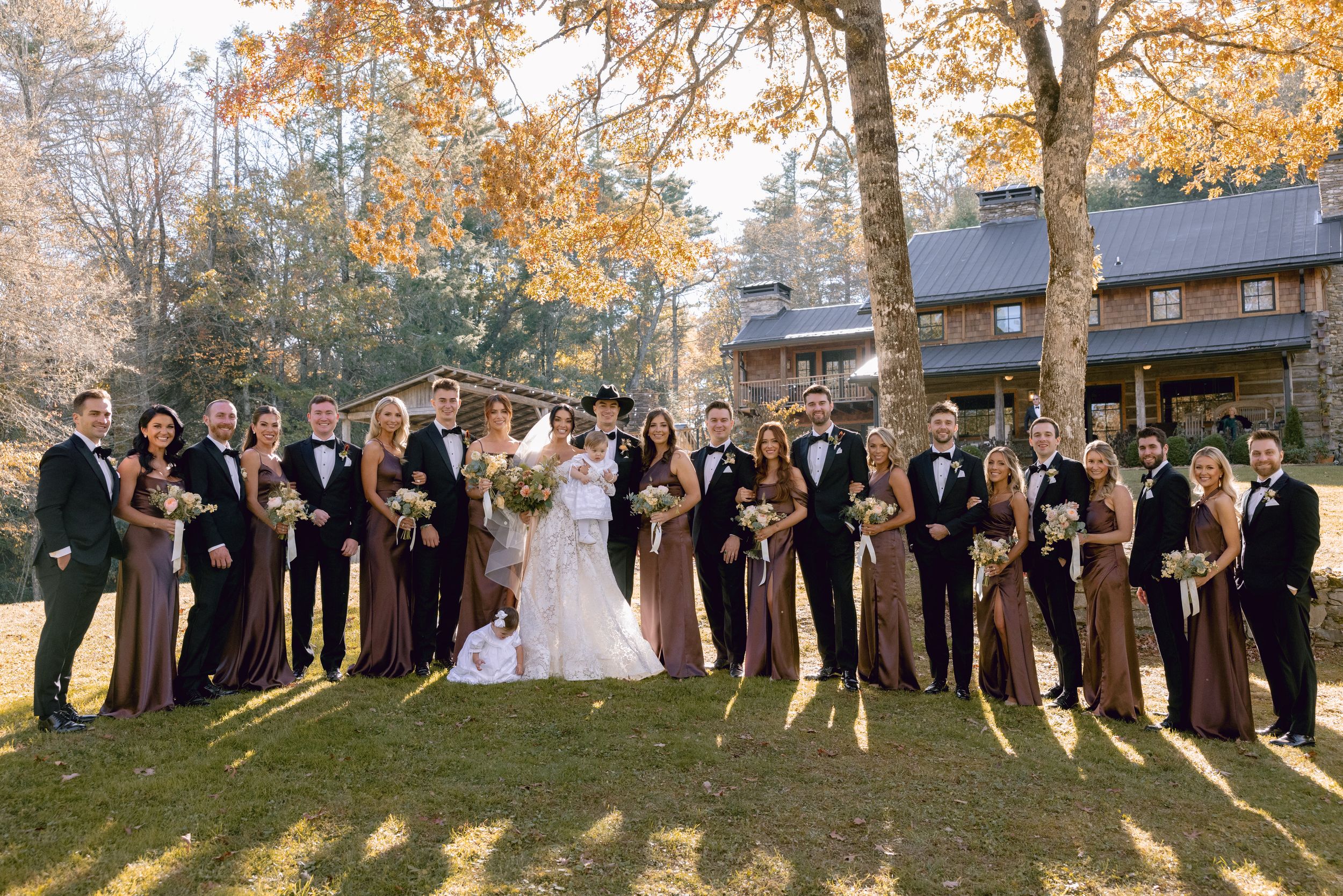 Wedding party stands on autumn lawn in formal attire with fall foliage and rustic lodge in background.