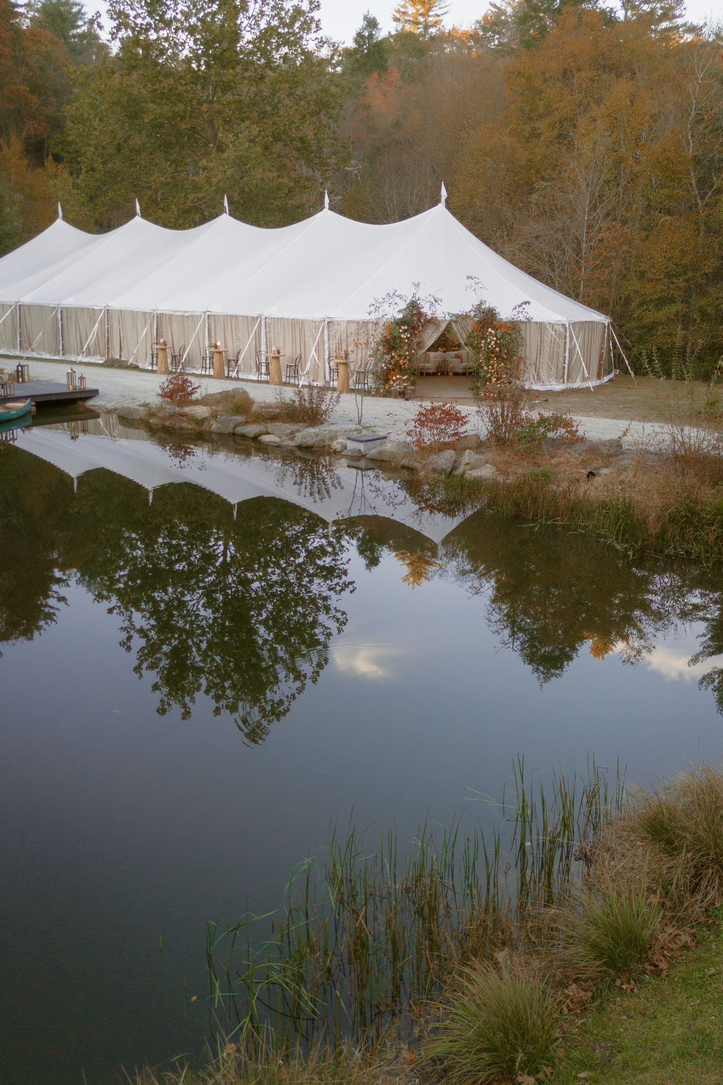 A large white wedding tent reflected in a serene pond surrounded by autumn trees in a natural outdoor setting.