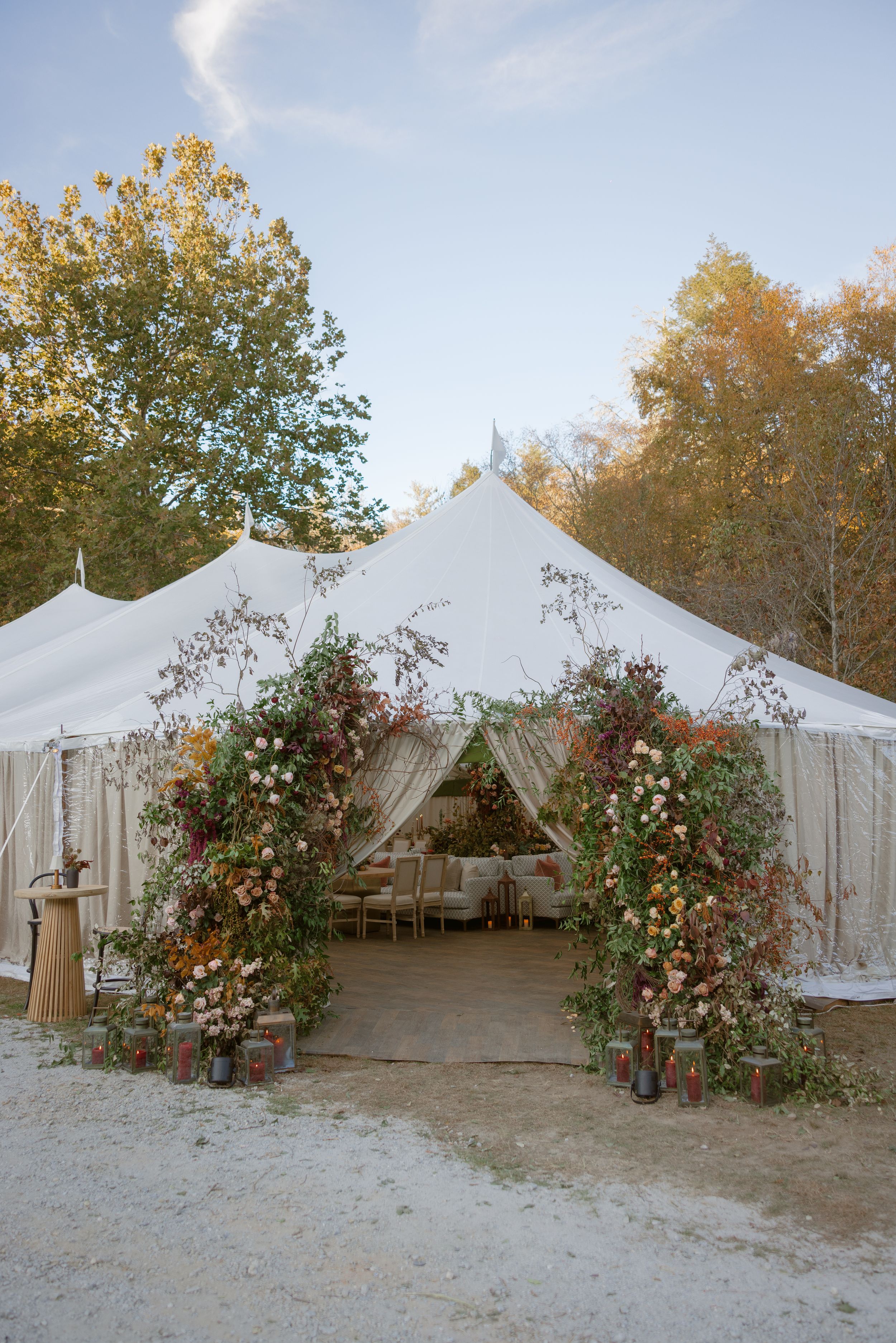 A white wedding tent entrance decorated with floral arrangements and fall foliage creates an elegant outdoor venue.