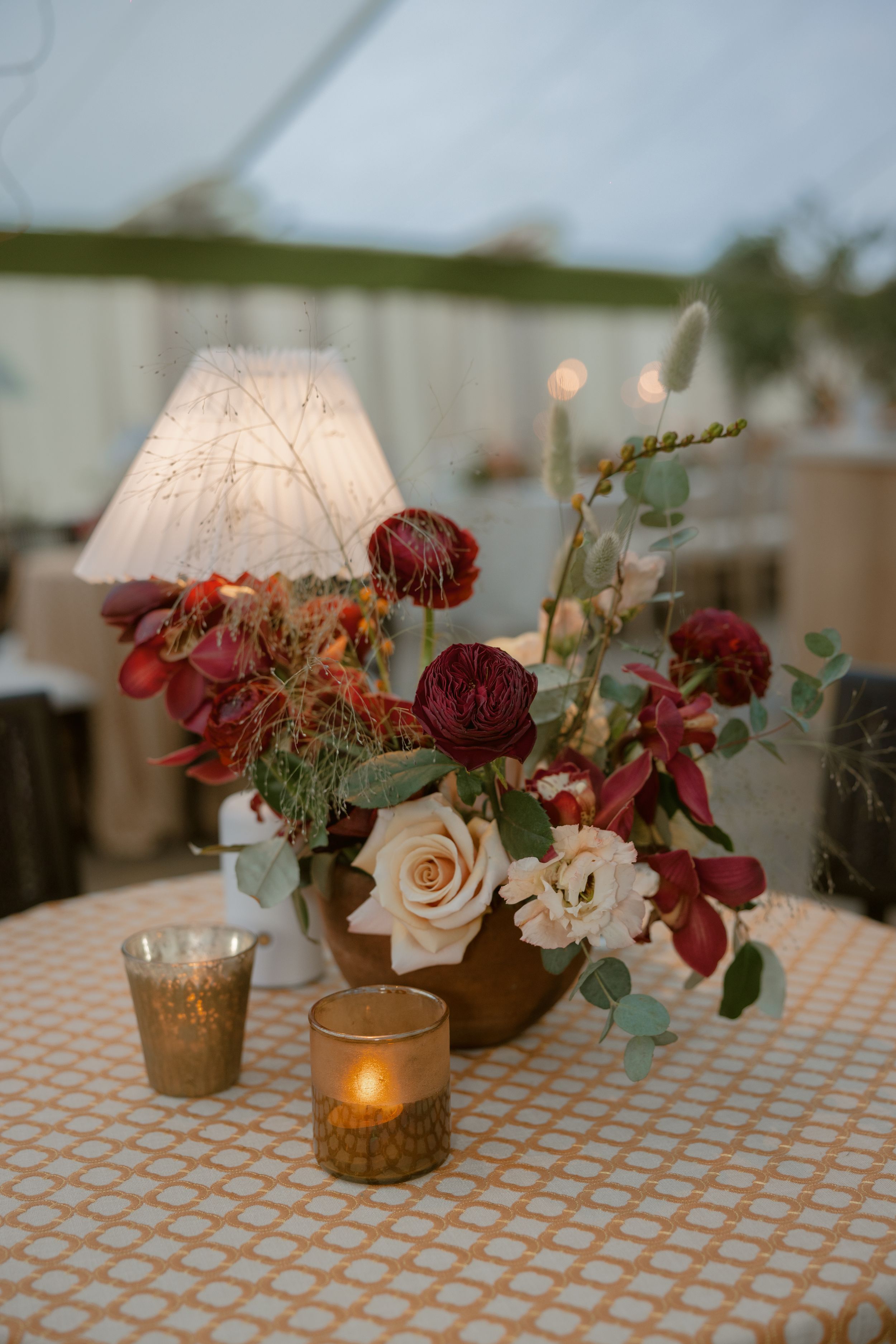 Elegant floral centerpiece with burgundy and cream roses, table lamp, and glowing candles on a checkered tablecloth.