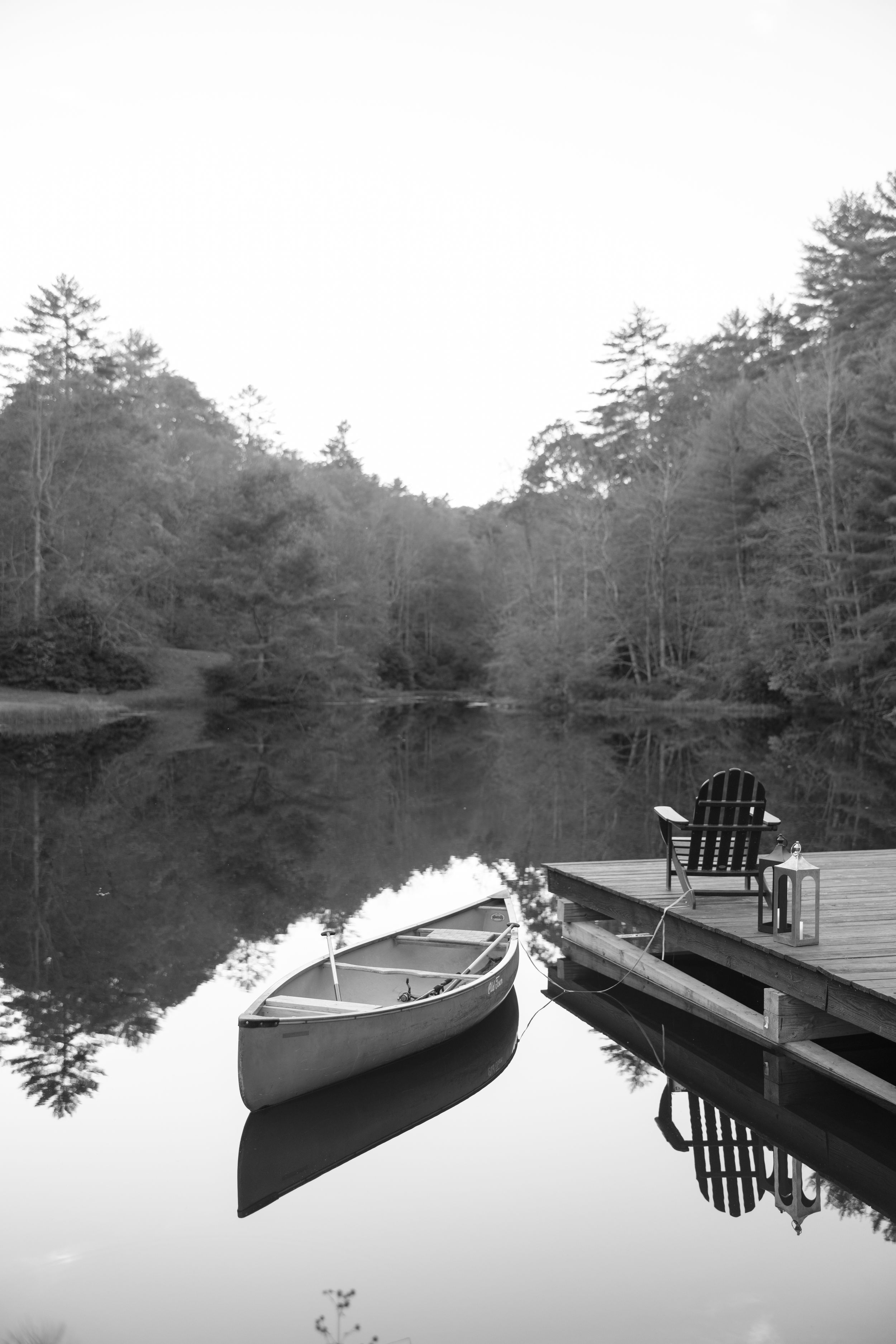 Canoe docked at wooden pier with Adirondack chair overlooking serene lake reflections in black and white.