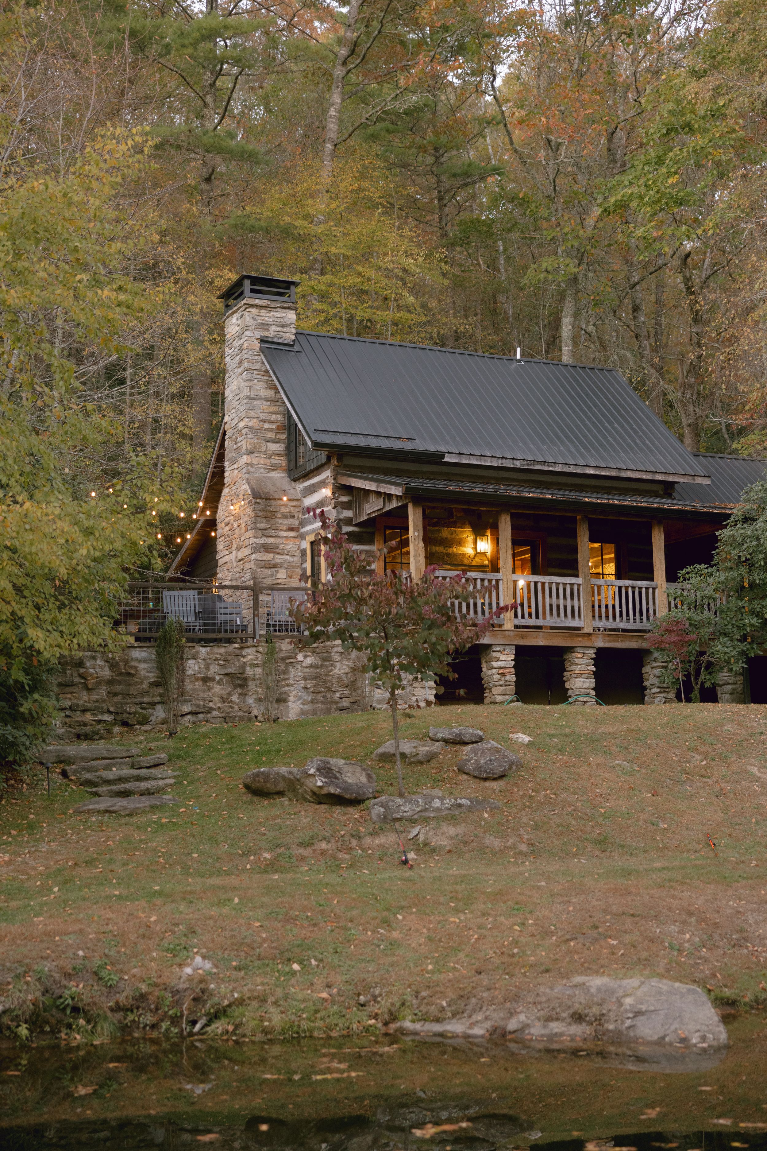 Rustic log cabin with stone chimney and front porch illuminated at dusk in autumn forest setting.