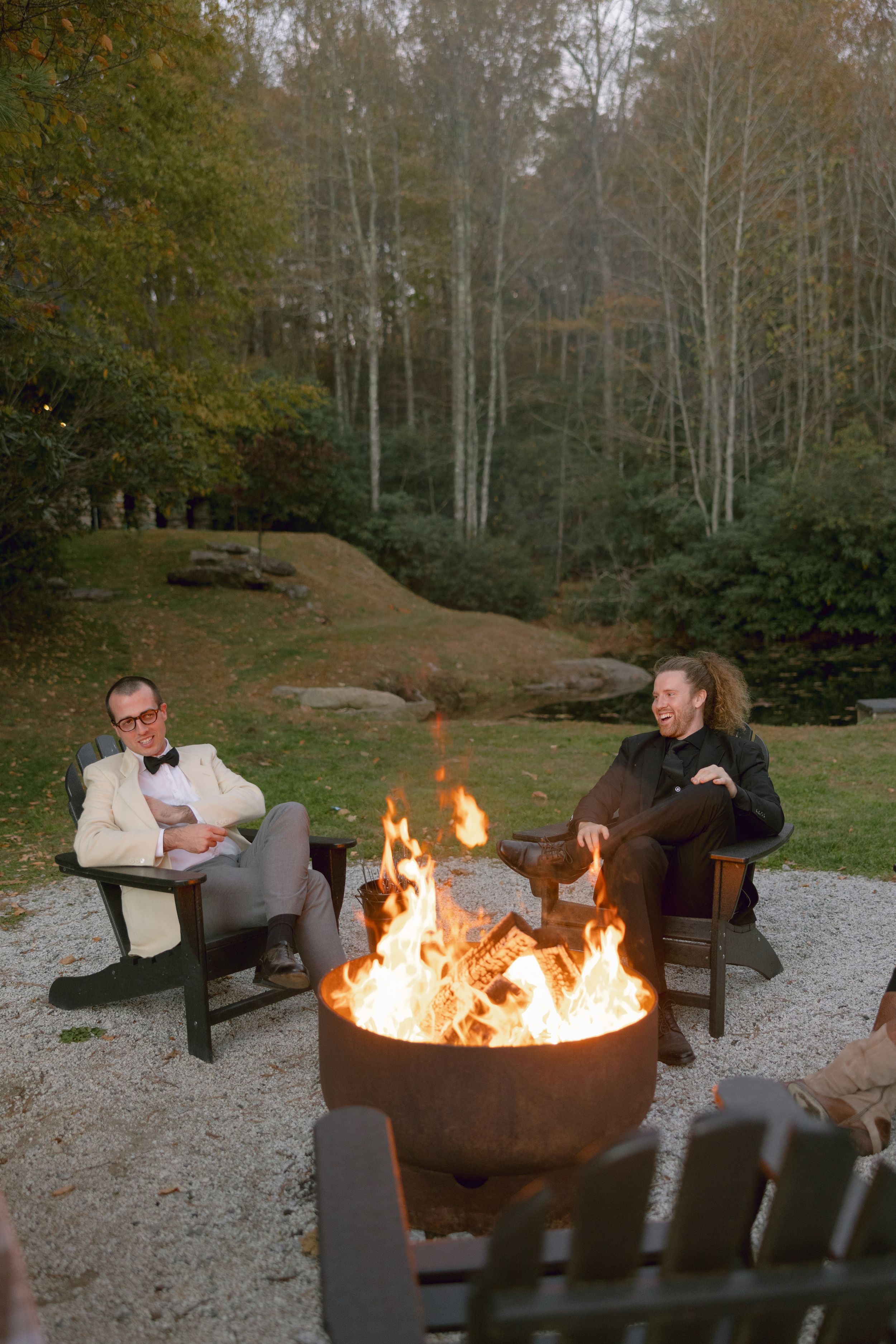 Two people relax in Adirondack chairs around a large fire pit on a graveled area with autumn woods in the background.