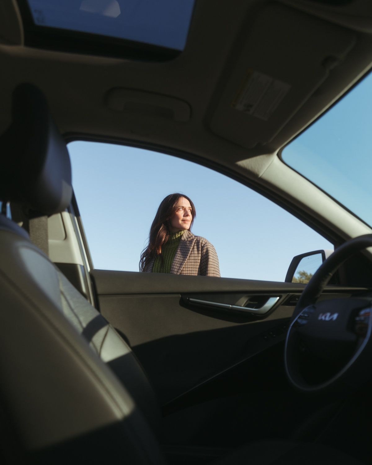 View through car window showing a person in a brown jacket smiling outside against a blue sky background.