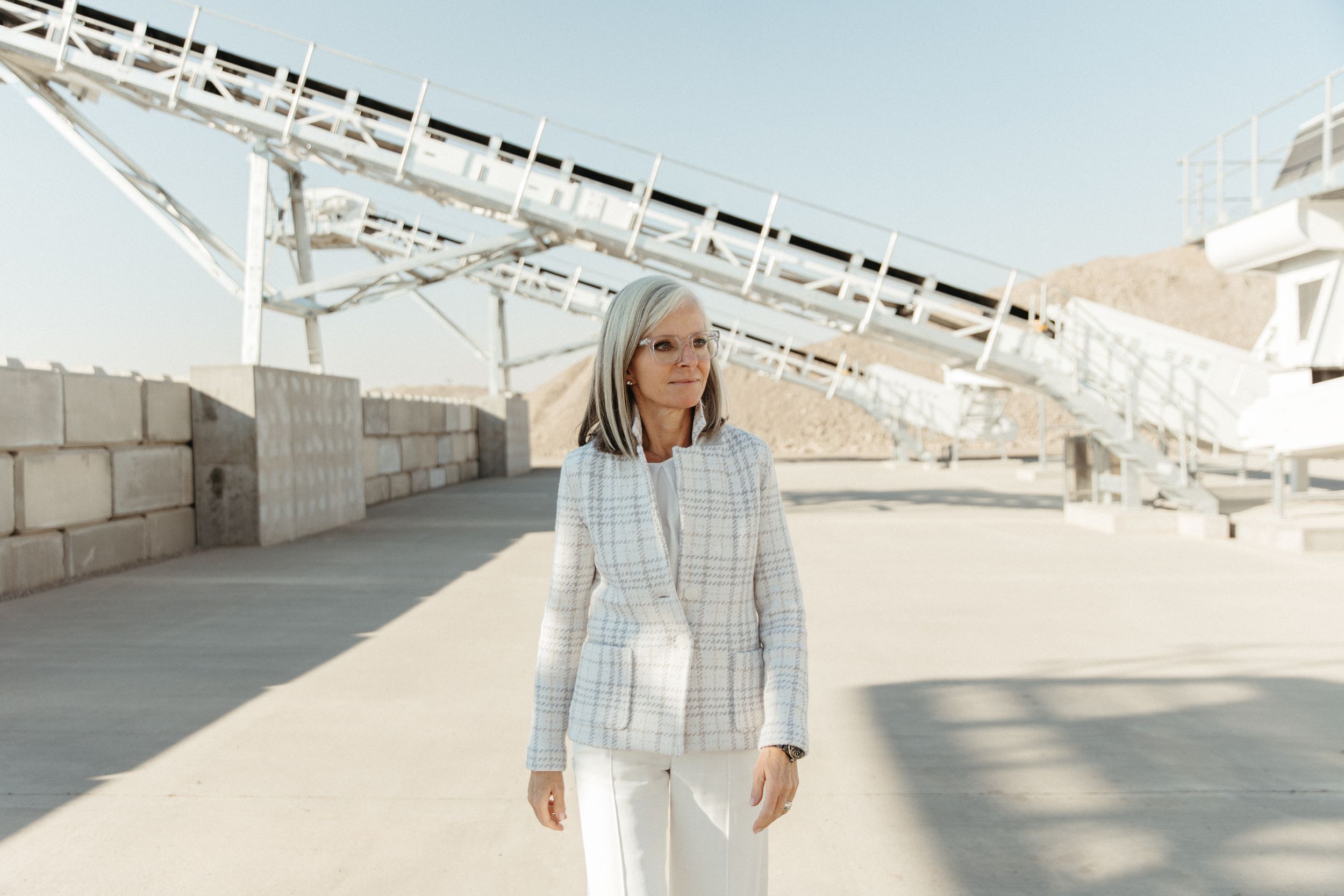 Person in a light plaid jacket and white pants stands near an architectural structure on a sunny day.