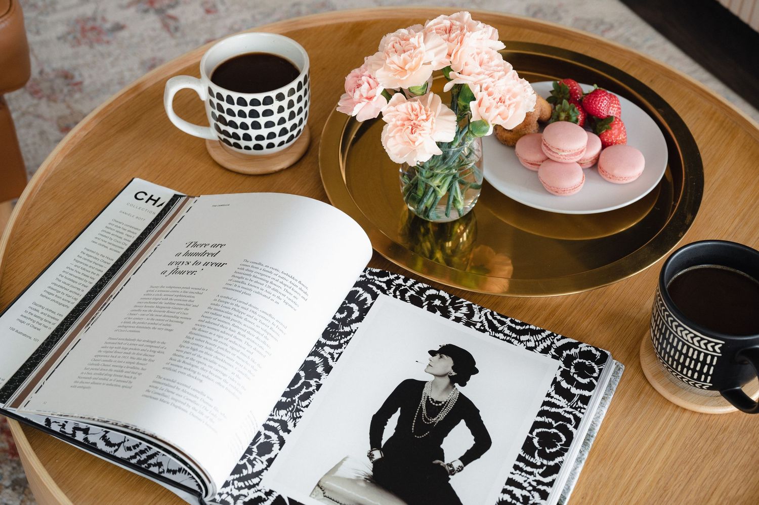 A wooden table displays an open magazine, pink roses in a vase, and coffee mugs with black and white geometric patterns.