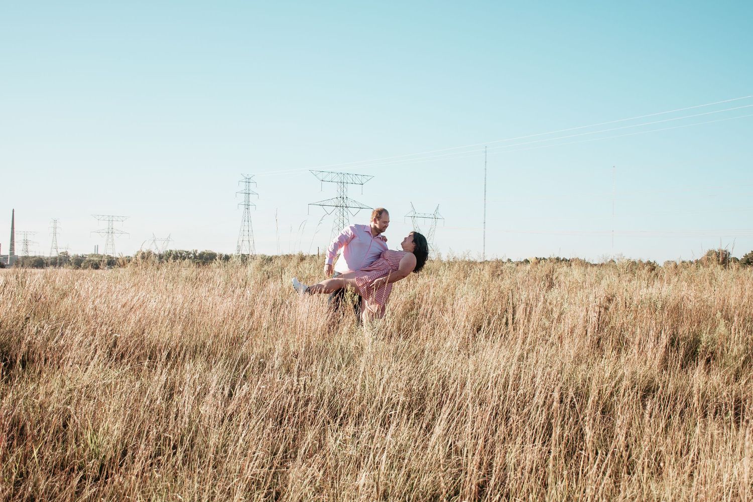 Couple standing together in golden prairie field with power lines in background.