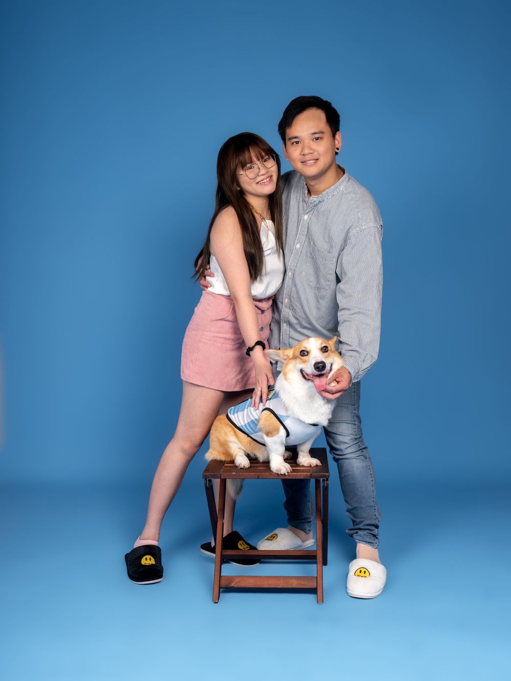 A couple poses with their small dog wearing a sailor outfit on a wooden stool against a bright blue background.