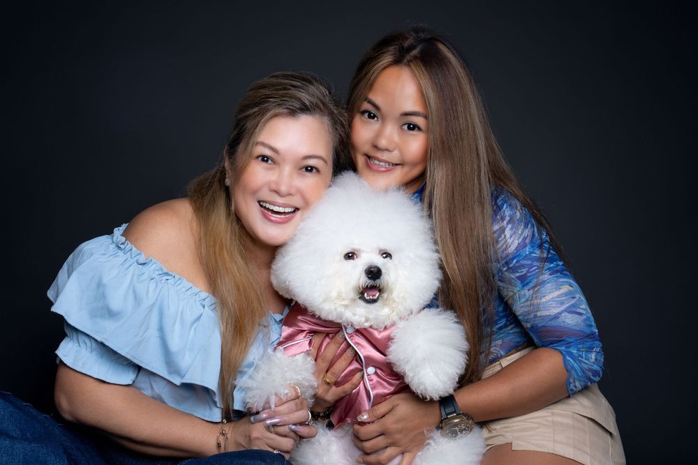 Two people wearing blue tops pose with a fluffy white Bichon Frise dog wearing a pink bowtie against dark background.