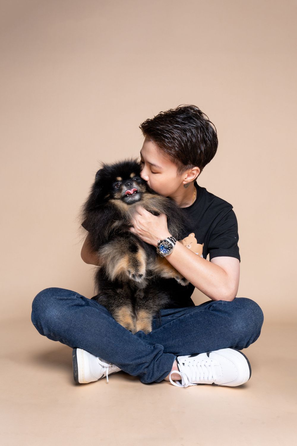 A person in black shirt and jeans sits cross-legged while cuddling an adorable black and tan Pomeranian dog against beige backdrop.
