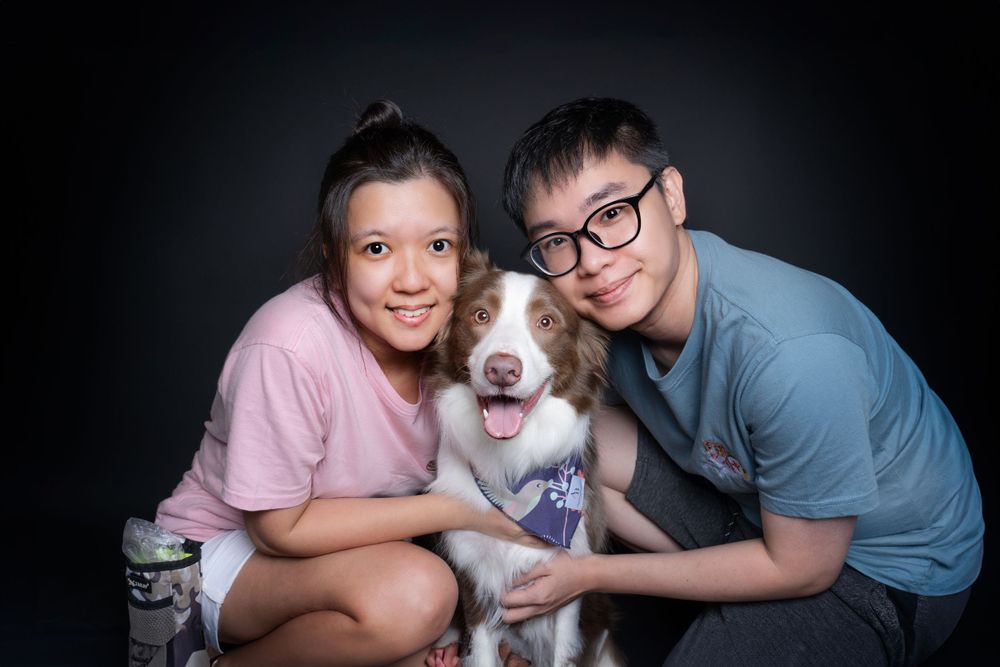 Border Collie sits between two people for family portrait against dark background.