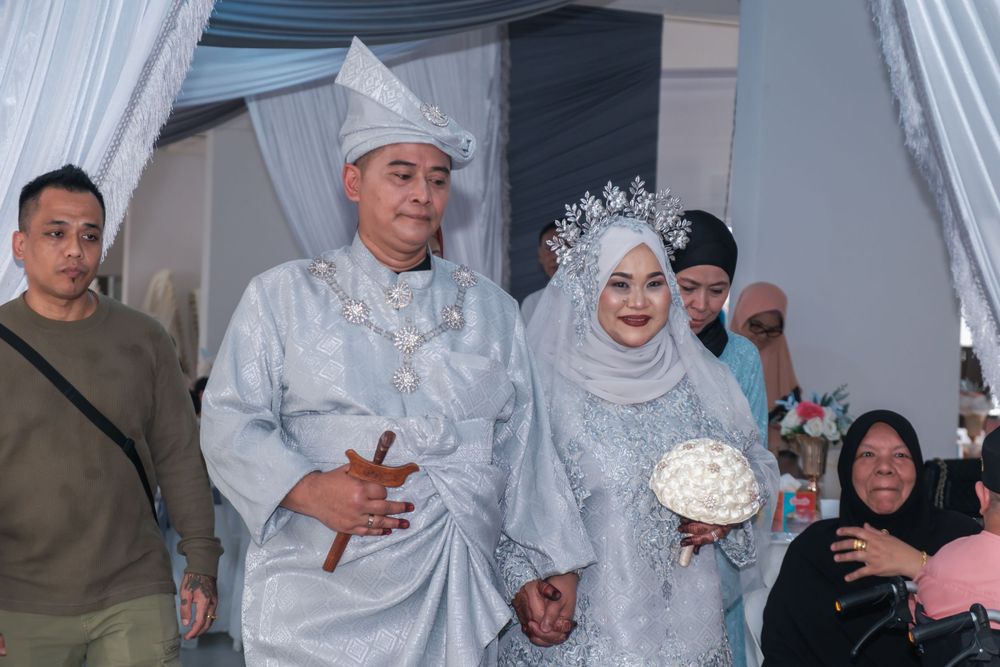 A wedding couple in matching silver traditional outfits walking under white draped decorations.
