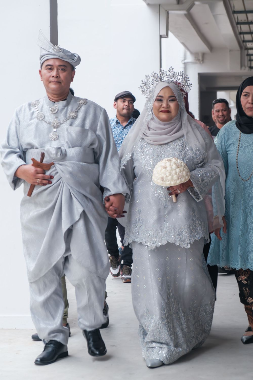 A couple in elegant silver Malay wedding attire walking together during their ceremony.