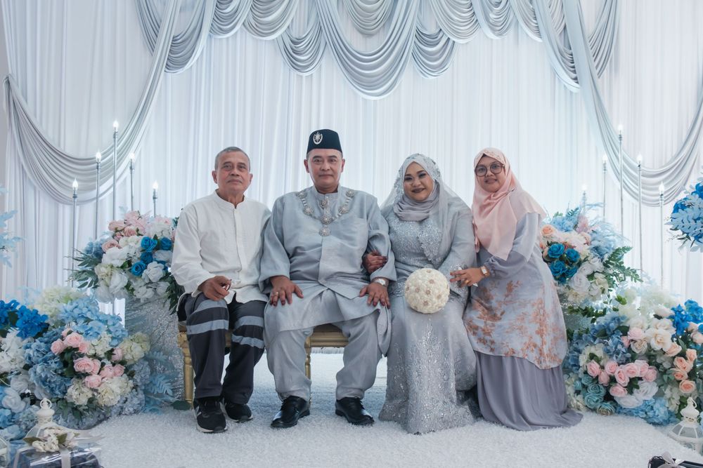 Wedding party in traditional Malay attire posing against floral backdrop with blue and white decor.