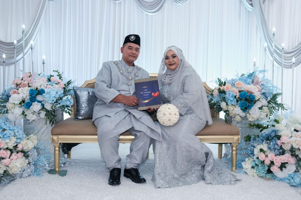 Newlyweds in silver and white attire posing together on decorated bench with blue flowers.
