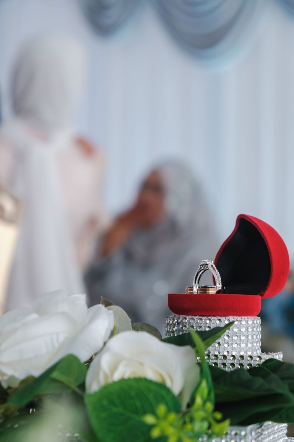 Close-up of a diamond ring in a red velvet box with white roses in the foreground.