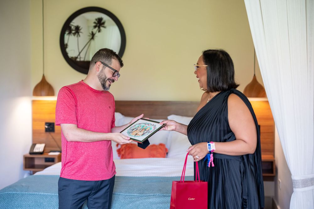 Two people exchanging a framed item in casual bedroom setting with palm tree wall art.