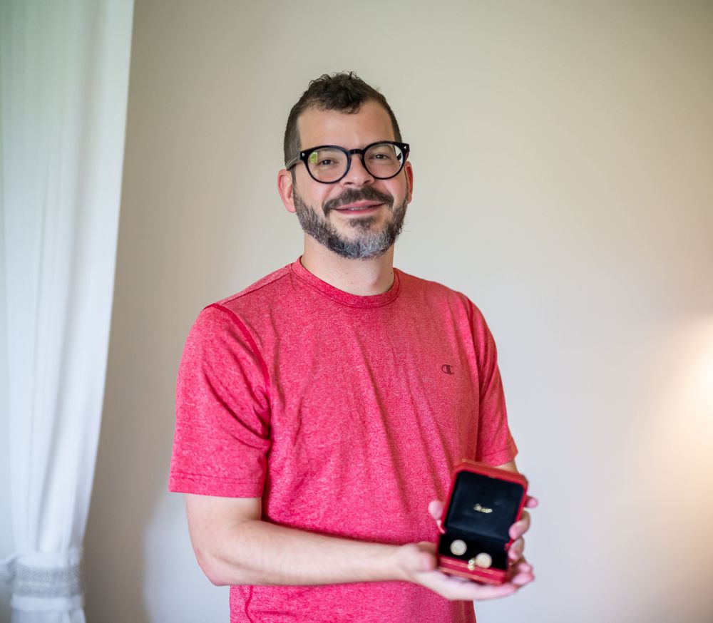 A person in a red t-shirt holding a small ring box while smiling near a window.
