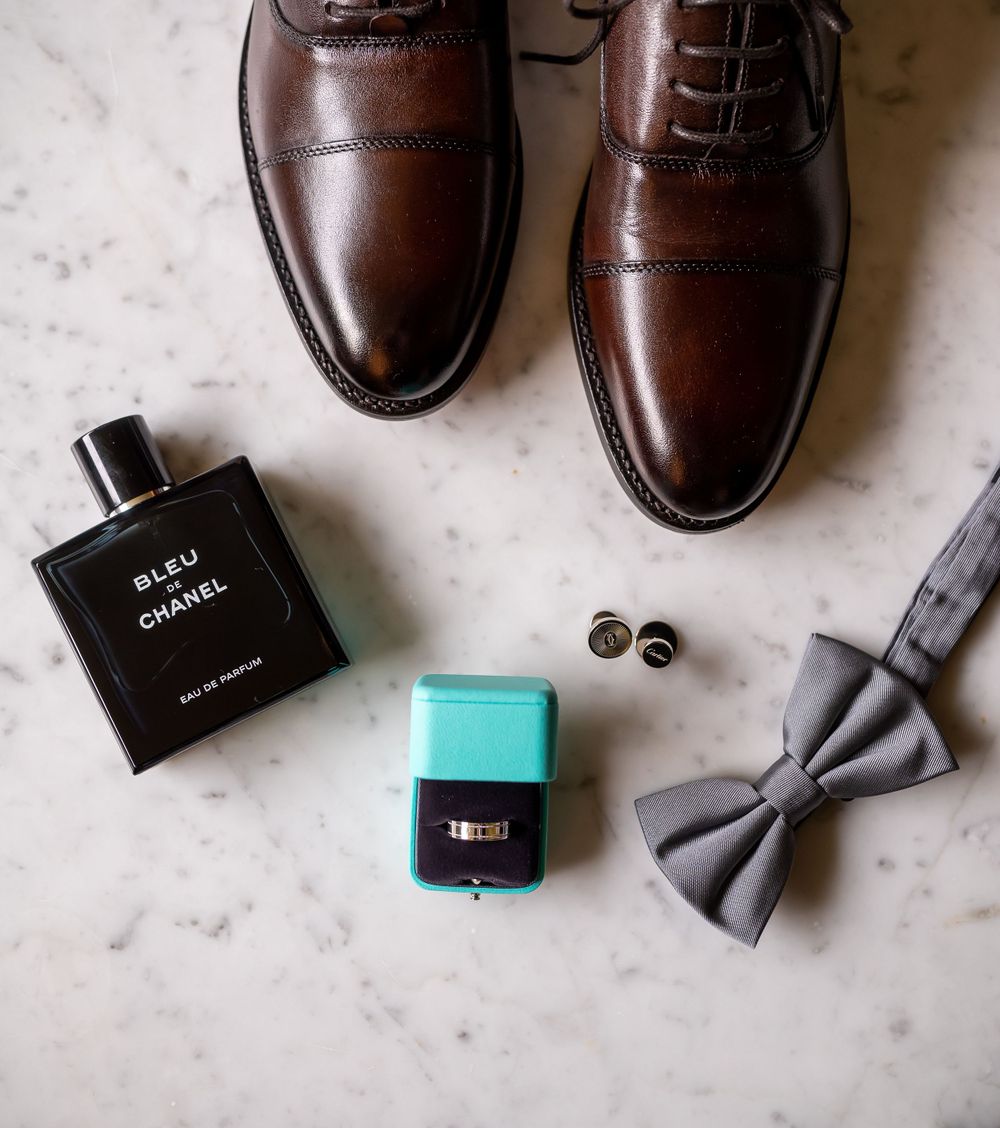 Wedding accessories including brown dress shoes, cologne, bow tie and ring box on marble surface.