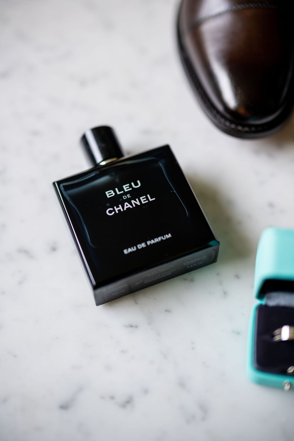 Chanel Bleu cologne bottle and turquoise ring box next to leather dress shoe on marble surface.