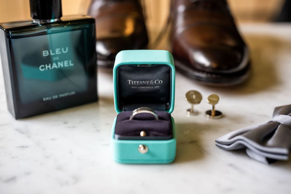 Wedding accessories arranged with Chanel cologne, ring box and dress shoes on marble counter.