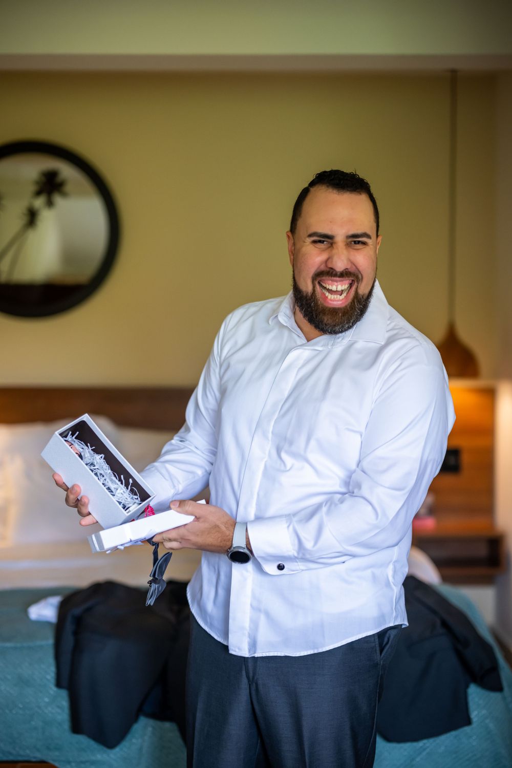 Person in white dress shirt holding gift box and smiling in hotel room.