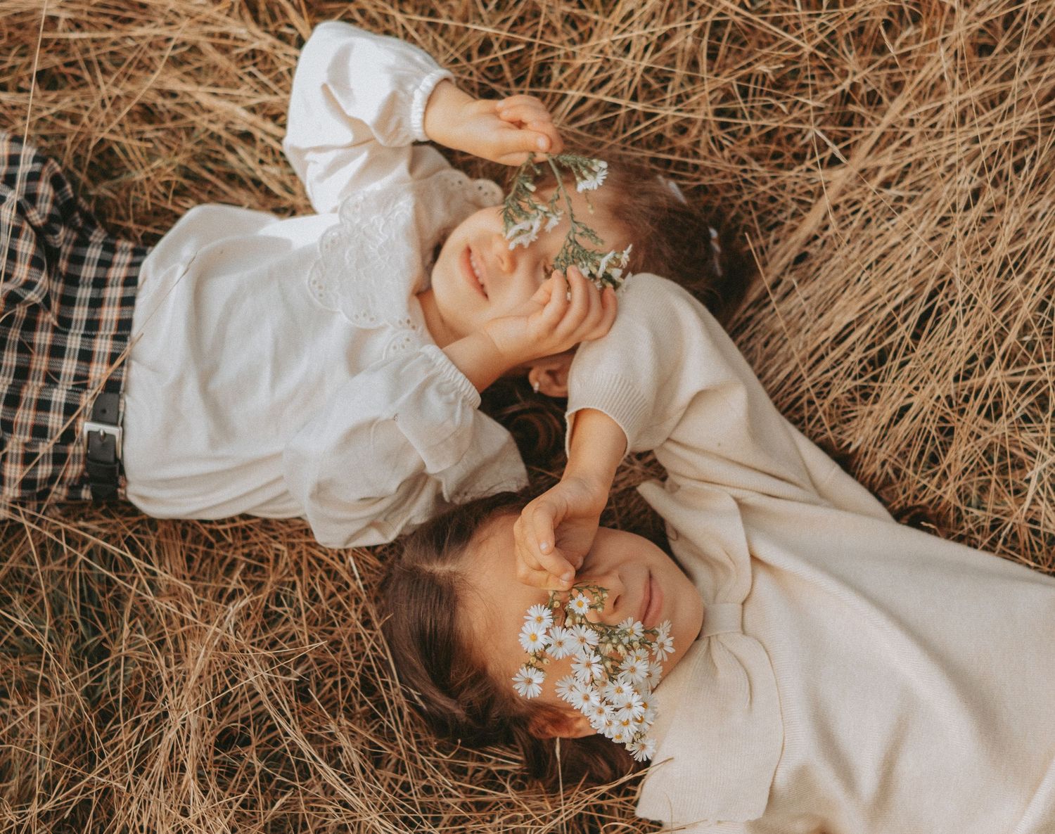 Two people in white dresses lay in golden dried grass together in warm sunlight creating a dreamy rustic scene.