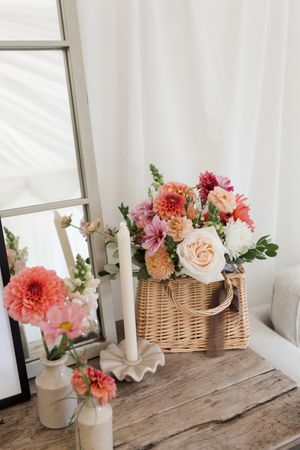Rustic table display with coral and pink floral arrangements in woven baskets and white ceramic vases near windows.