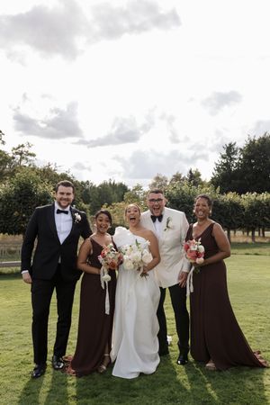 Wedding party in brown and white attire poses together on a sunny lawn with trees in background.
